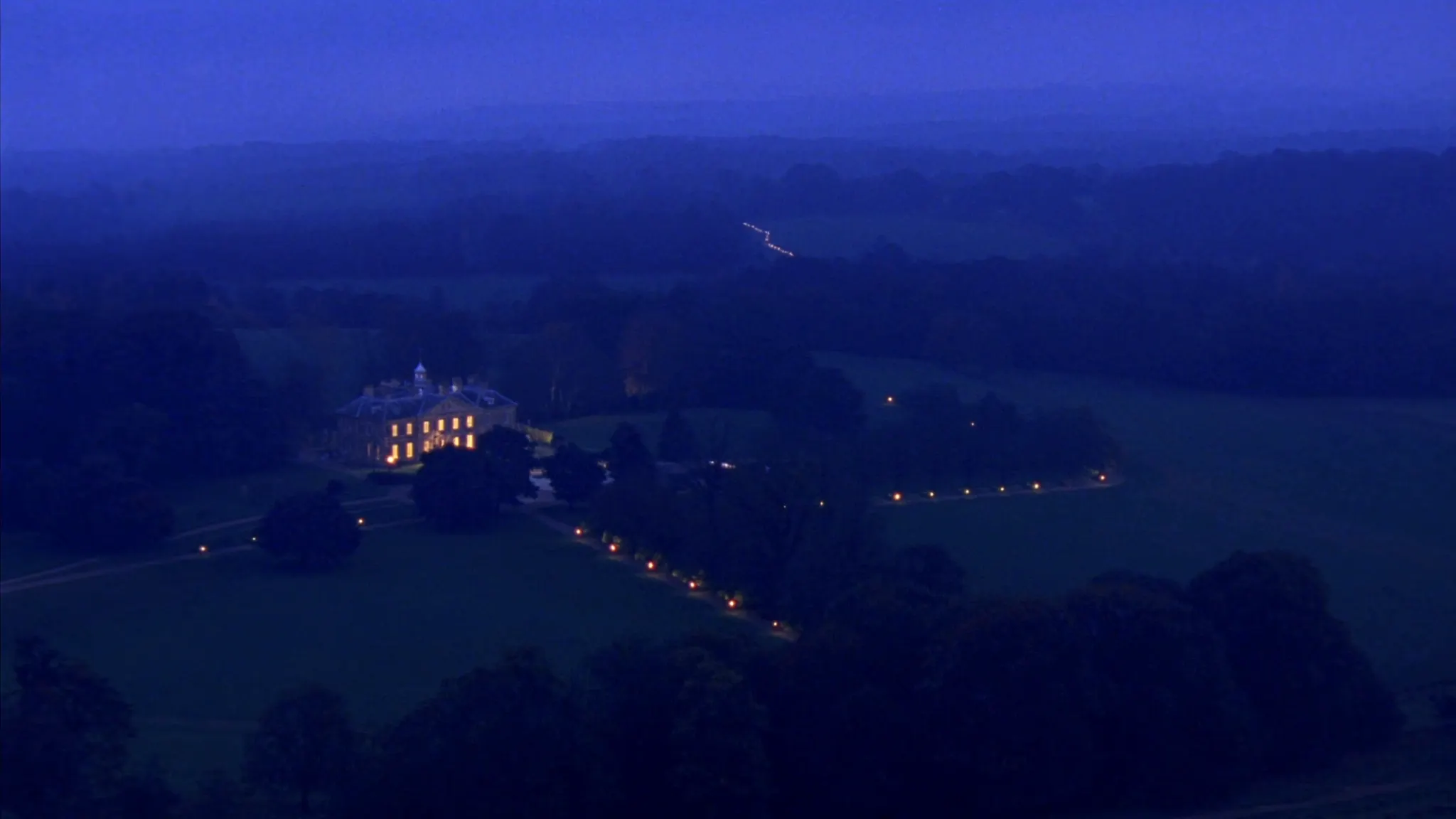 A lamp-lit drive winding to a country house at dusk
