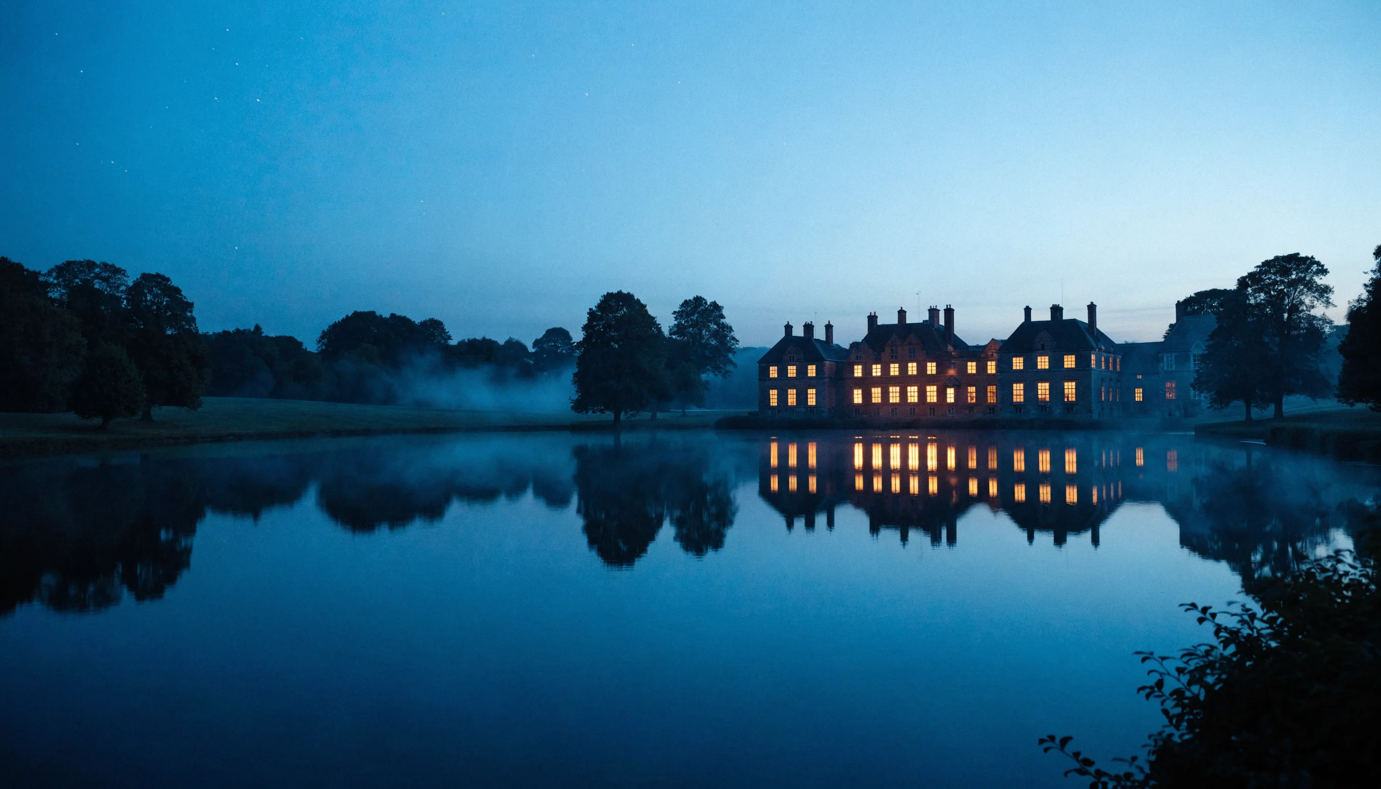 A grand English manor at blue hour, every window glowing warm, reflected in a still lake